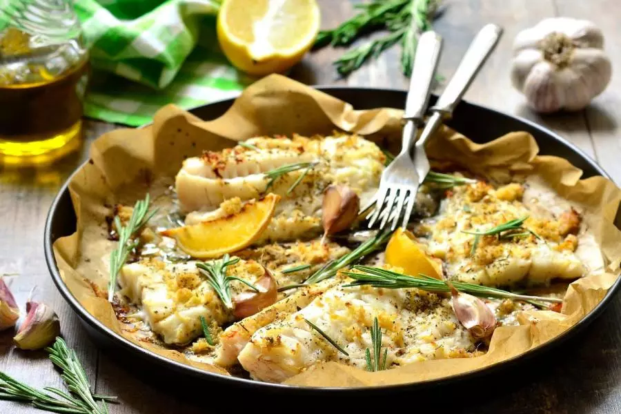 Baked cod fillets on parchment in a pan, topped with rosemary, garlic, and lemon wedges, with olive oil, fresh herbs, and a halved lemon in the background.