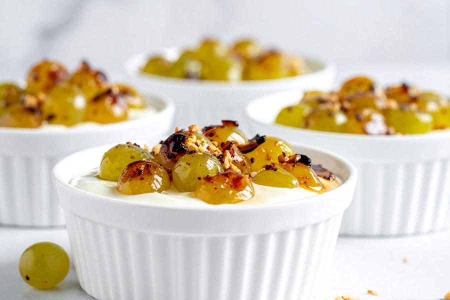 White ramekins filled with yogurt topped with roasted green grapes and toasted nuts on a bright white background.