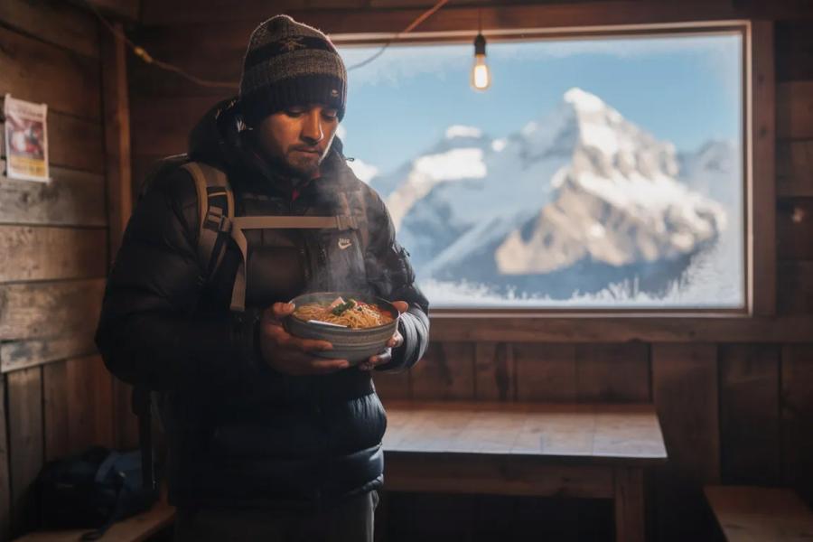 A trekker sits inside a cozy teahouse, holding a steaming bowl of thukpa, a popular Nepali dish made with noodles, vegetables, and spices, while snowy mountains are visible through the window, showcasing the breathtaking landscape of Nepal. The scene captures the essence of Nepali cuisine and the warmth of local hospitality amidst the majestic Himalayas.