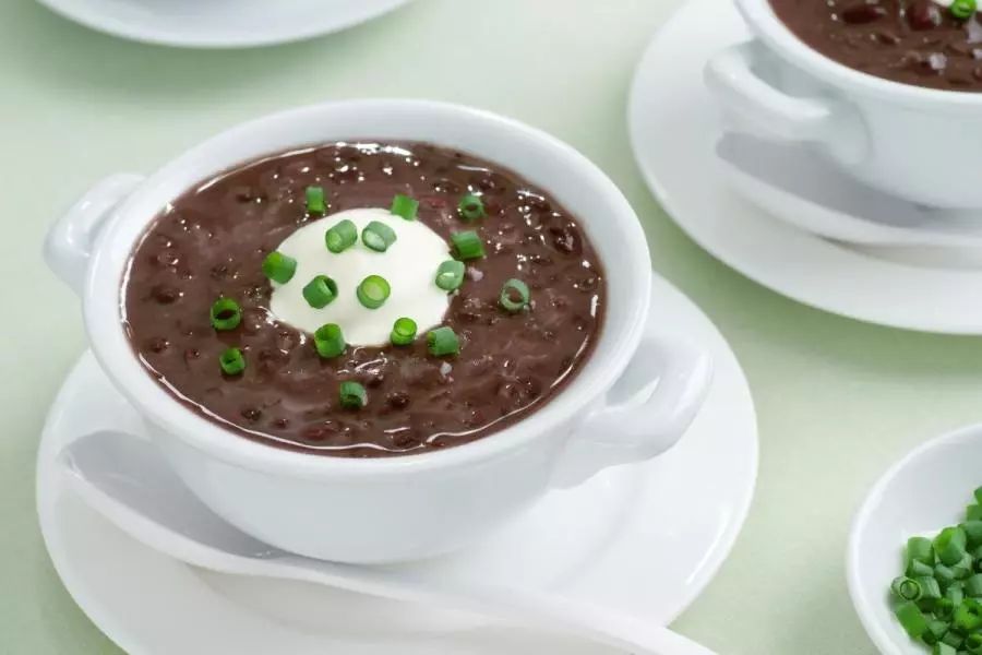 White handled bowl of thick chili black bean soup topped with sour cream and green onions, served on a saucer with a soup spoon.