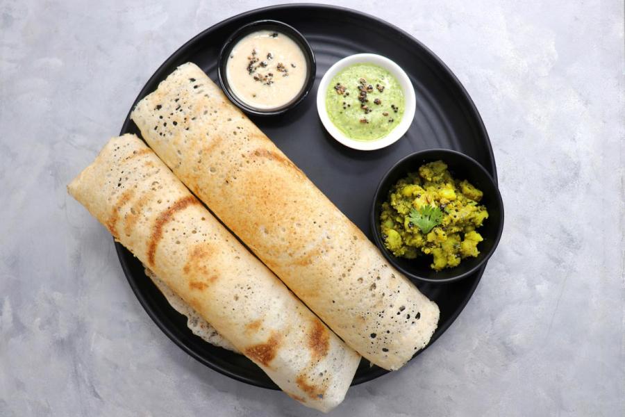 Two rolled masala dosas on a black plate, served with coconut chutney, green herb chutney, and a bowl of spiced potato filling, viewed from above.