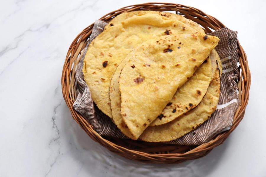Stack of freshly made whole-wheat rotis with charred spots, served in a round wicker basket lined with a linen cloth on a white marble surface.