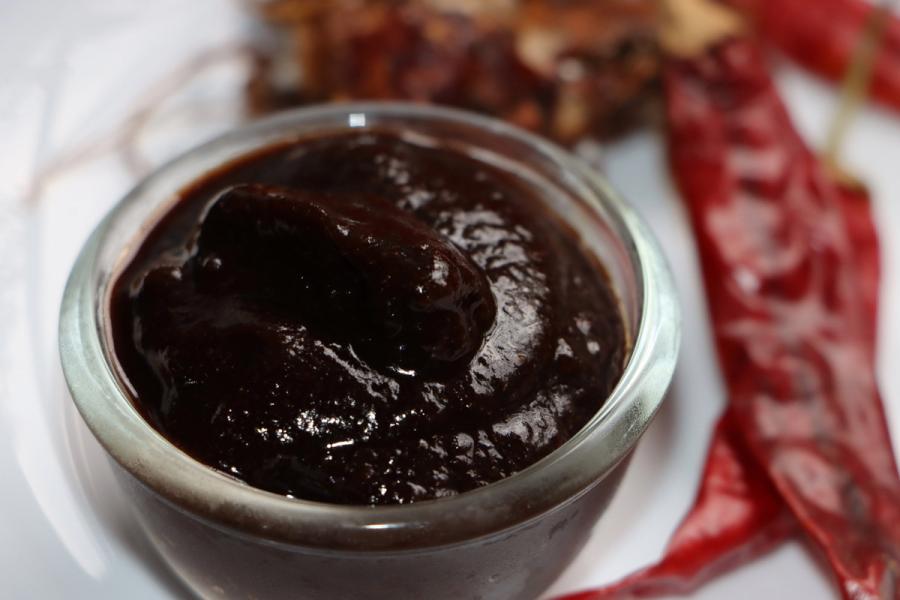 A small glass bowl filled with thick, dark tamarind chutney, with dried red chili peppers and tamarind pods blurred in the background.