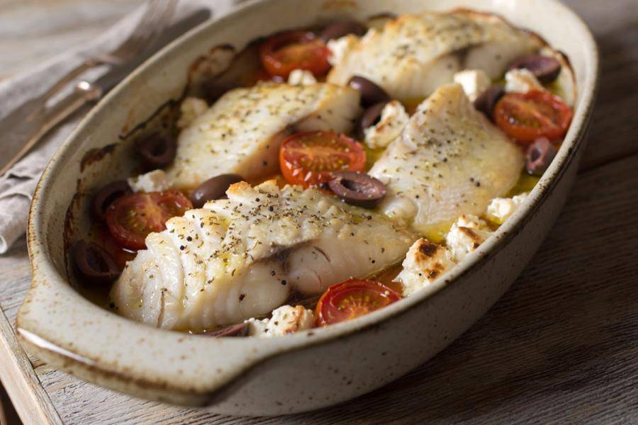 Baked white fish fillets with cherry tomatoes, olives, and feta in an oval baking dish - synthetic image.