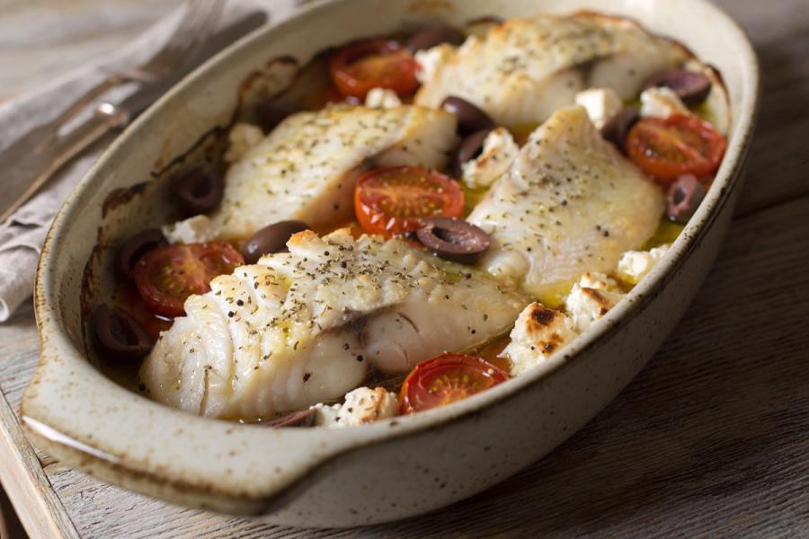 Baked white fish fillets with cherry tomatoes, olives, and feta in an oval baking dish - synthetic image.