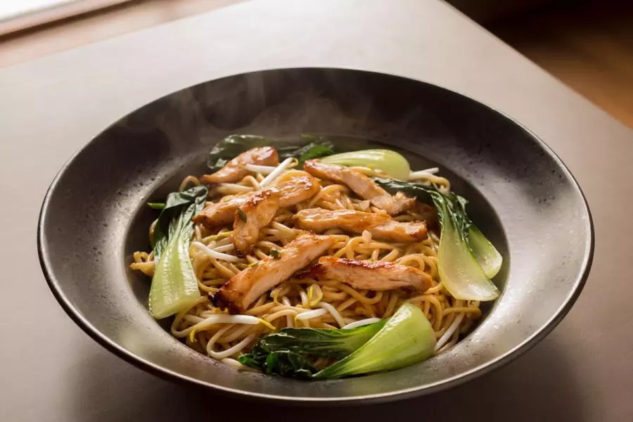 Steaming bowl of wok-fried noodles topped with sliced glazed chicken, bok choi, and bean sprouts. This is a synthetic image.