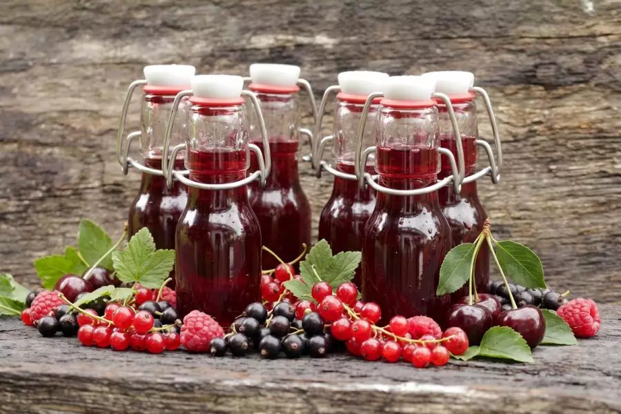 Six small swing-top glass bottles of deep red summer berry cordial on a rustic wooden surface, surrounded by fresh redcurrants, blackcurrants, raspberries, and cherries.