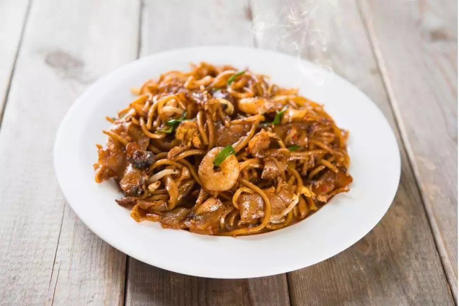 A plate of char kway teow — stir-fried flat rice noodles with shrimp, egg, bean sprouts and scallions, glistening with dark soy sauce and lightly charred, served on a white plate on a wooden table, with steam rising.
