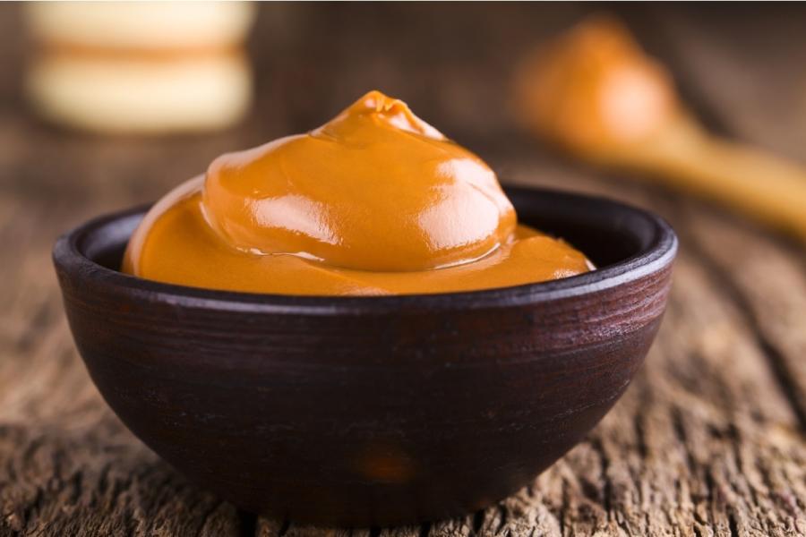 A dark ceramic bowl filled with glossy dulce de leche, with a wooden spoon and a jar visible in the blurred background.