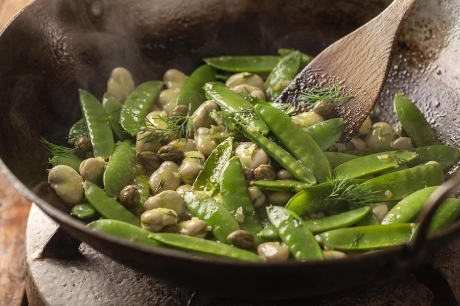 A wok filled with sautéed fava beans, sugar snap peas and capers, garnished with fresh dill, held with a wooden spatula and gently steaming.