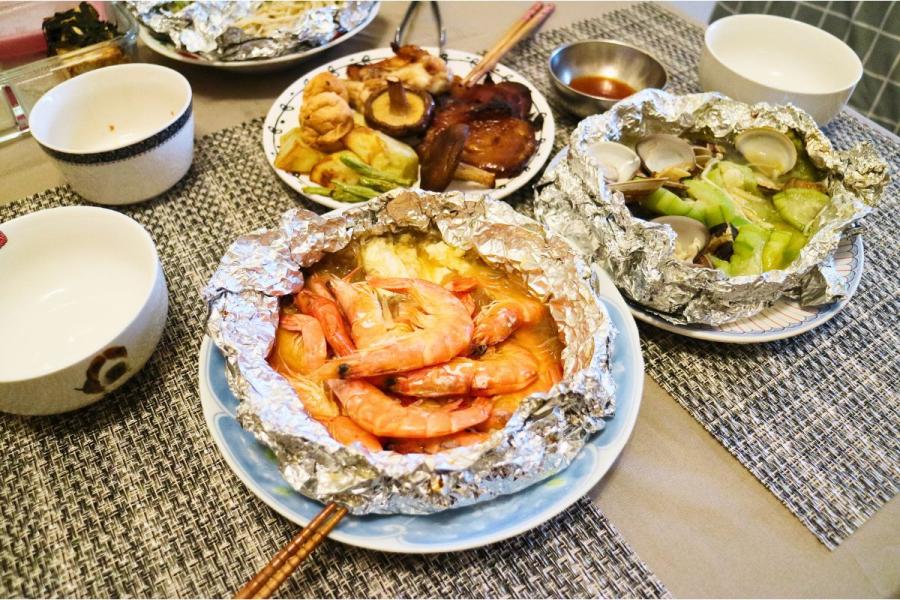 Several foil packet meals on a table, including glazed grilled meats and vegetables, shrimp in a savory broth, and clams with vegetables in silver foil, served alongside small bowls and chopsticks.