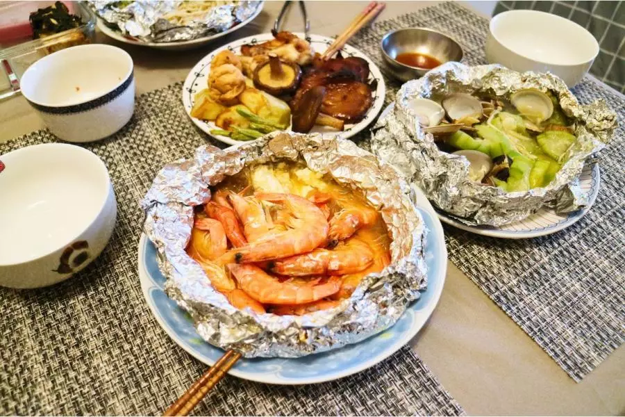 Several foil packet meals on a table, including glazed grilled meats and vegetables, shrimp in a savory broth, and clams with vegetables in silver foil, served alongside small bowls and chopsticks.