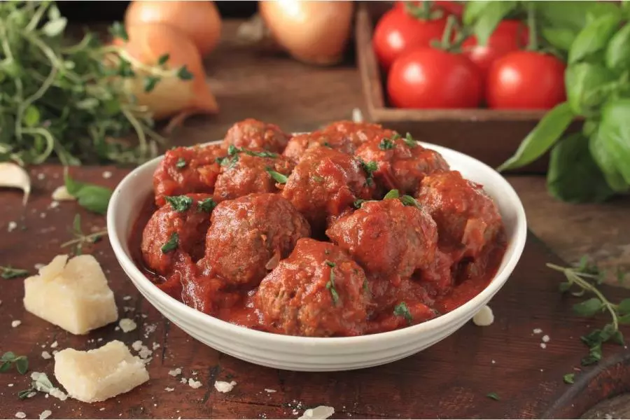 A white bowl of Italian meatballs in rich tomato sauce, sprinkled with fresh herbs, with basil, tomatoes, onions and chunks of Parmesan on a rustic wooden table.