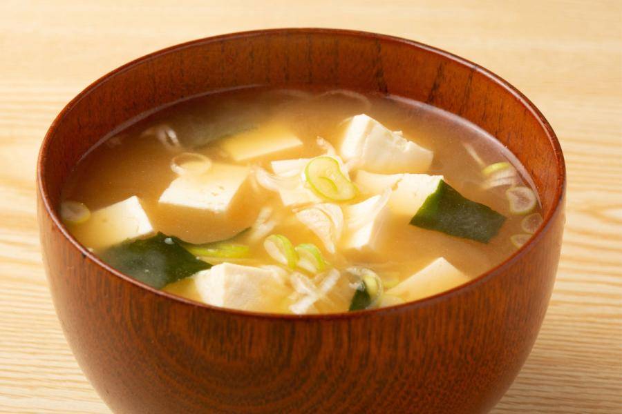 A wooden bowl of miso soup filled with cubes of soft tofu, dark green wakame seaweed, and sliced green onions in a golden broth, on a light wooden table.