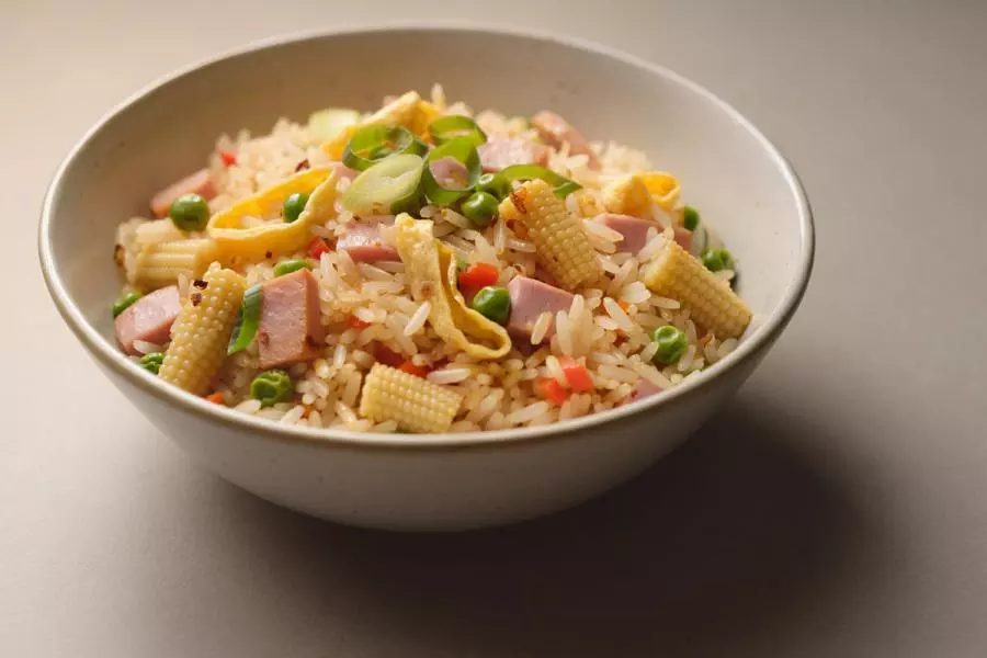 A bowl of fried rice with diced ham, egg ribbons, baby corn, peas, carrots and scallions, on a warm beige background, synthetic image.
