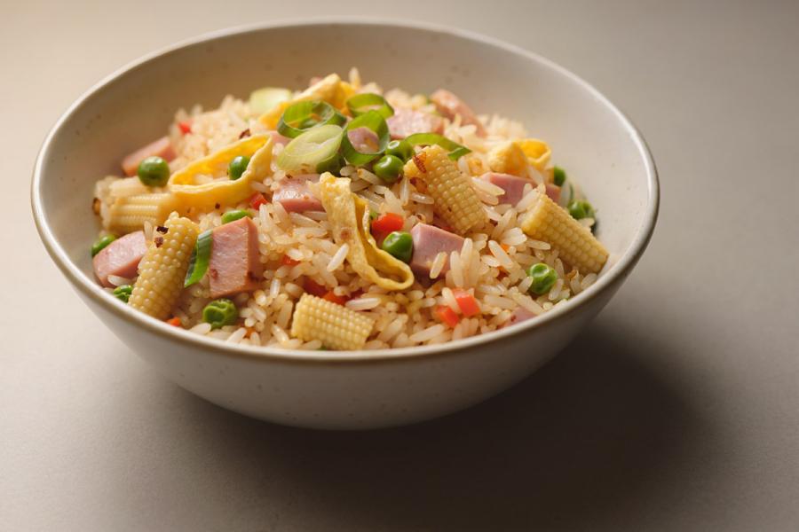 A bowl of fried rice with diced ham, egg ribbons, baby corn, peas, carrots and scallions, on a warm beige background, synthetic image.