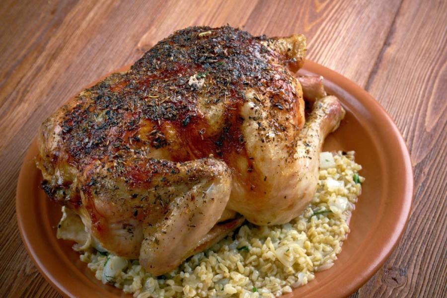 A whole roasted squab, seasoned with a dark herb crust, served on a bed of freekeh pilaf on a terracotta plate.