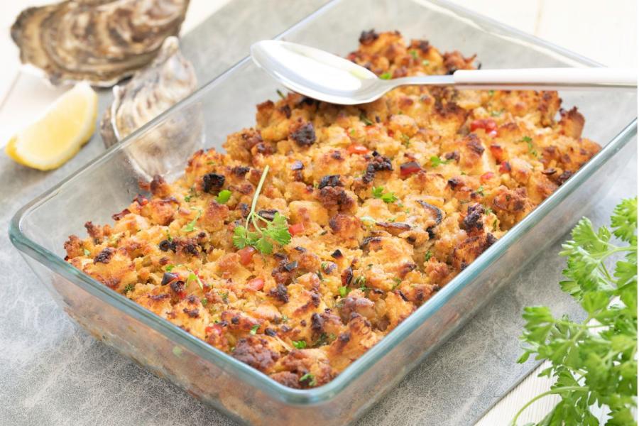 Golden-baked oyster and cornbread stuffing in a clear glass baking dish, flecked with red pepper and parsley, with a serving spoon, oyster shells and a lemon wedge alongside