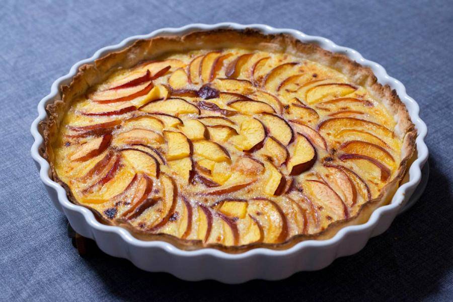 An apple tart in a white fluted ceramic dish, thin apple slices arranged in overlapping rings over a set custard filling, golden and caramelized at the edges, on a blue-gray linen cloth.
