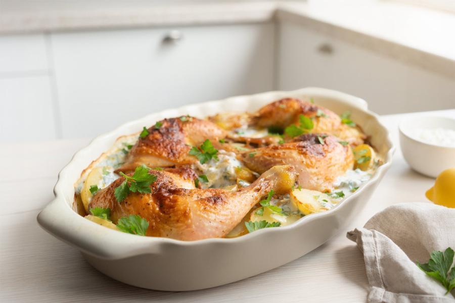 Baked chicken legs with sliced potatoes in a creamy lemon-yogurt sauce, garnished with fresh parsley, in a white oval baking dish.
