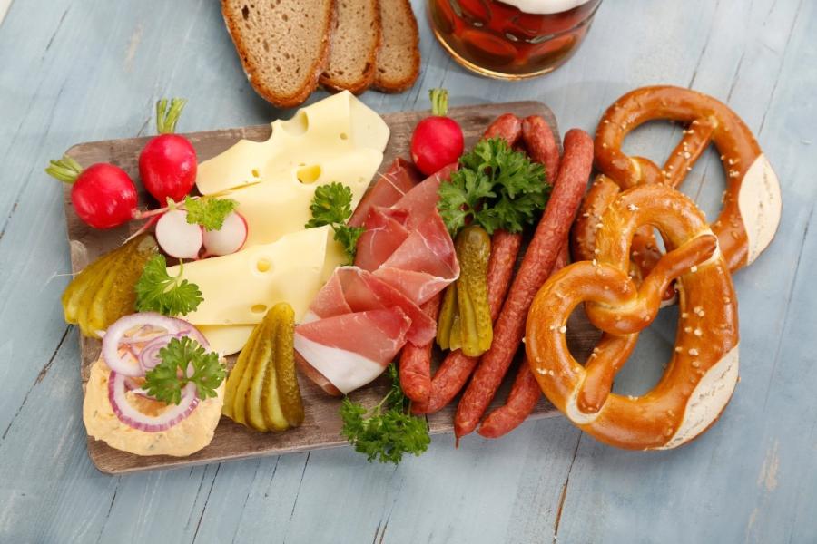Wooden board of Bavarian cold cuts — Emmental cheese, cured ham, thin smoked sausages, gherkins, radishes and Obatzda — with two pretzels, rye bread and a beer alongside