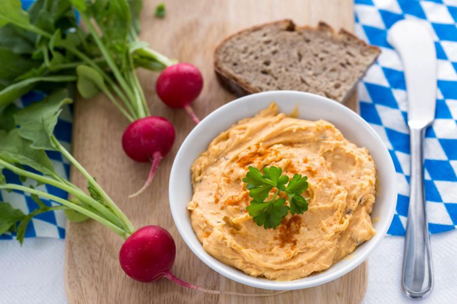 A bowl of creamy orange Obatzda dusted with paprika and topped with a parsley sprig, served with whole radishes and slices of dark rye bread on a blue-and-white checkered napkin.