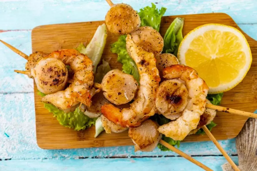 Grilled scallop and shrimp skewers on a wooden board with lettuce leaves and a lemon slice, dusted with black pepper.