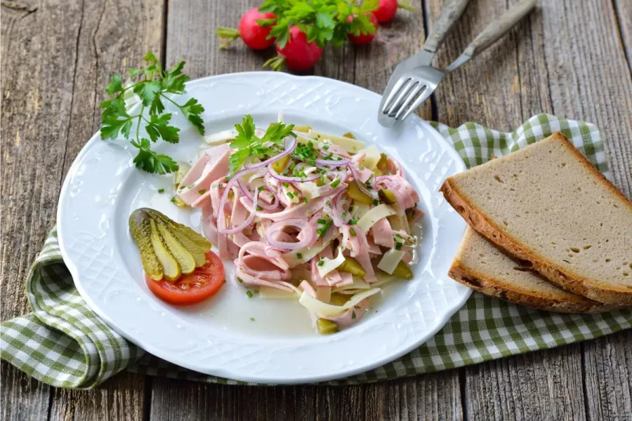 White plate of Bavarian sausage and cheese salad — strips of Lyoner sausage and Emmental with red onion rings, gherkin and chives — served with two slices of rye bread