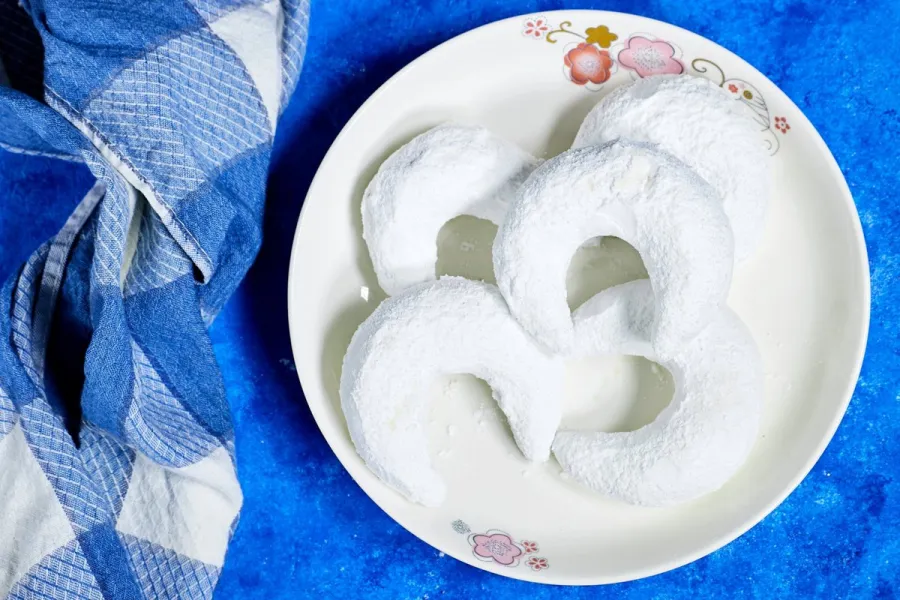 Crescent-shaped gazelle horn almond cookies heavily dusted with powdered sugar on a white floral-rimmed plate