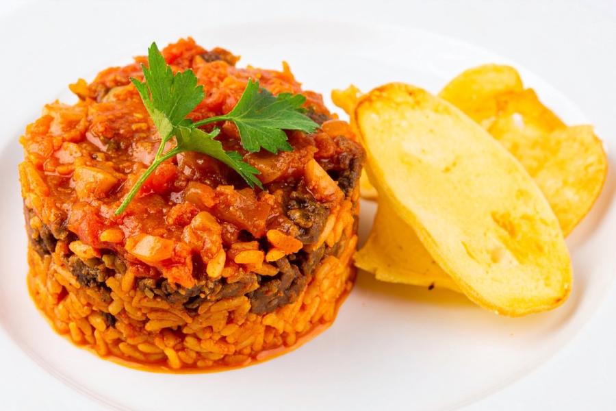 A molded round of spiced tomato-red rice topped with a savory sauce and a flat-leaf parsley sprig, with two golden fried potato slices alongside, on a white plate.