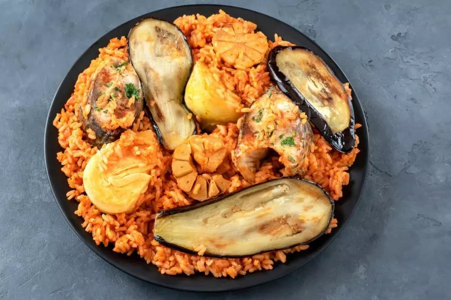 A black plate of ceebu jën: fish steaks on red tomato rice with fried eggplant wedges, cabbage, potato slices, and whole roasted garlic.
