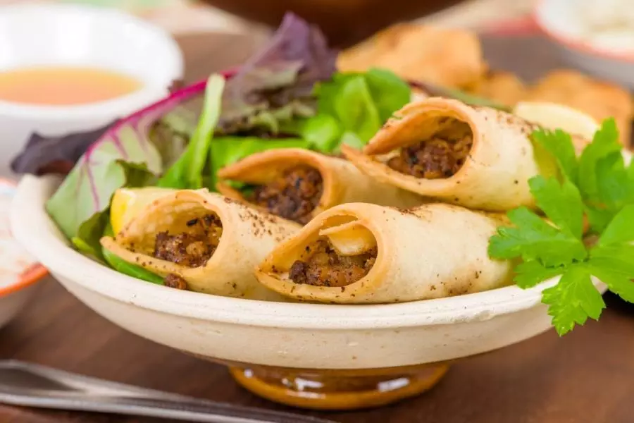 A bowl of kefta briouats — crisp cylindrical Moroccan pastry rolls filled with seasoned ground meat and dusted with cinnamon — served on salad leaves with lemon, fresh parsley, and a small dish of dipping sauce.