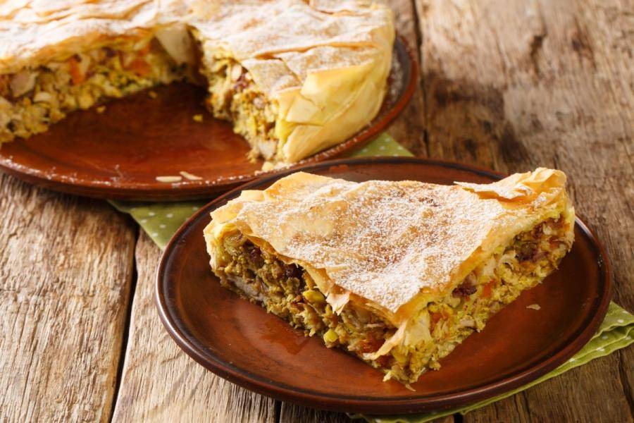 A wedge of Moroccan chicken pastilla on a rustic plate, its crisp golden phyllo crust dusted with powdered sugar, cut to reveal a spiced chicken, egg, and almond filling, with the rest of the round pie on a wooden table behind.