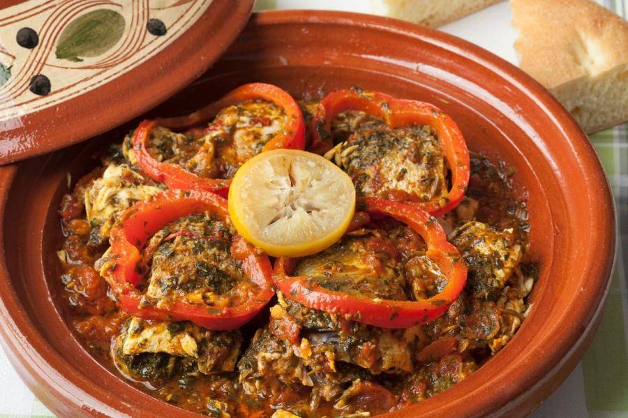 A Moroccan fish tagine in a clay dish: pieces of white fish in a chermoula-spiced tomato sauce, topped with rings of red bell pepper and half a lemon, with bread alongside.