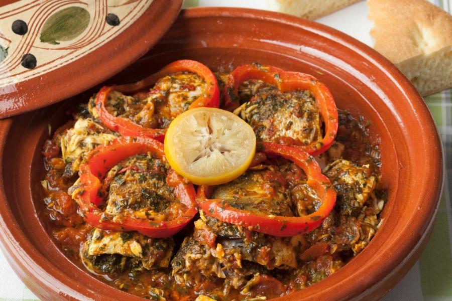 A Moroccan fish tagine in a clay dish: pieces of white fish in a chermoula-spiced tomato sauce, topped with rings of red bell pepper and half a lemon, with bread alongside.
