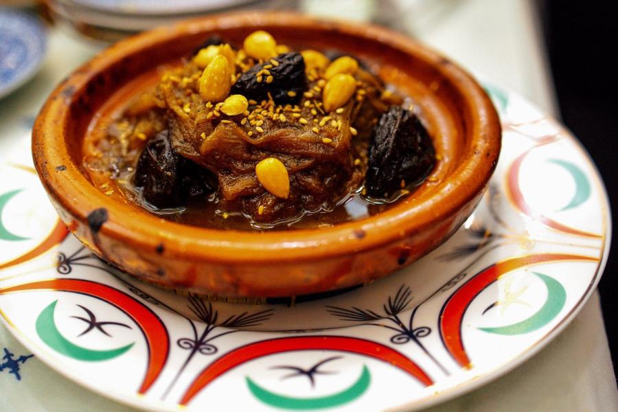 A Moroccan lamb and prune tagine (mrouzia) in a traditional clay tagine dish: dark, glossy slow-cooked lamb with whole prunes, topped with fried blanched almonds and sesame seeds, on a decorative patterned plate.