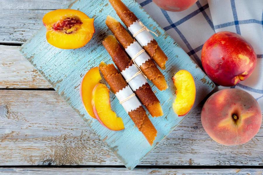 Rolled strips of peach fruit leather tied with paper and string on a pale-blue board, with fresh whole peaches, a halved peach and slices