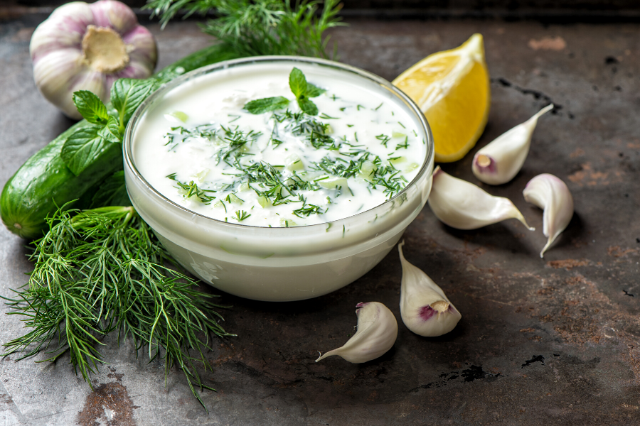 A bowl with tzatziki, a Greek yogurt sauce, surrounded by the ingredients required to prepare this sauce.