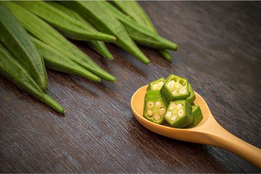 Fresh okra, a few whole pods and some chopped okra in a wooden spoon.