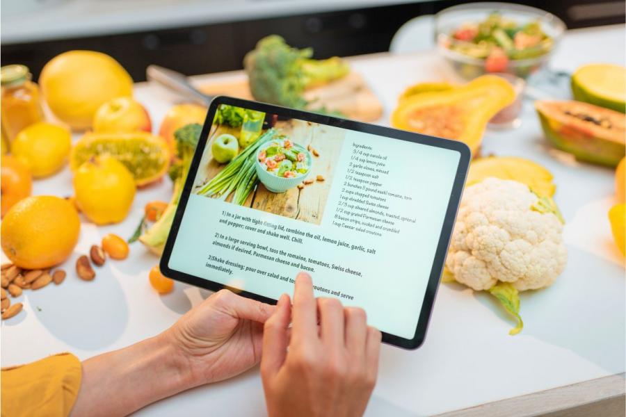 A tablet showing a recipe on screen and the table with ingredients to make it in the background.